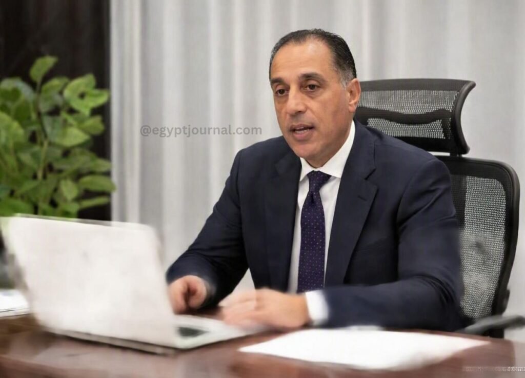 Man in a dark suit and purple tie sits at a desk with an open laptop, speaking or presenting in an office setting.