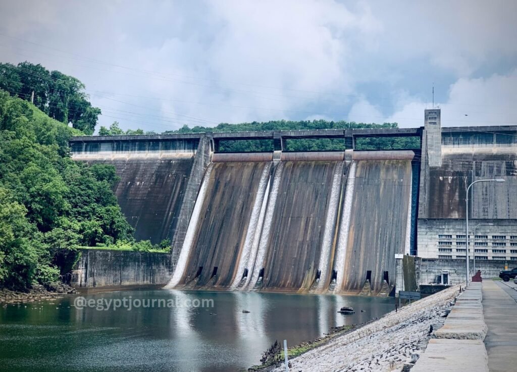 Concrete dam with several spillways releasing water into a calm pool, flanked by green trees and a walkway on the right under a cloudy sky.