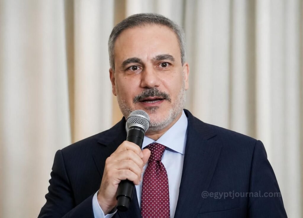Man in a dark suit and red tie speaking into a handheld microphone at a formal event in front of light curtains